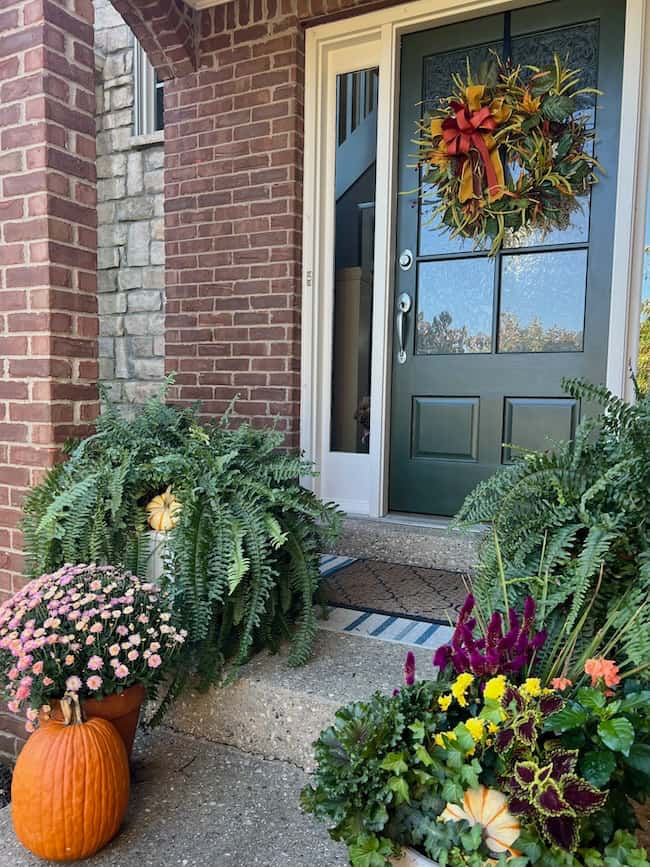 A classic home front porch decorated for fall features a dark green front door, a vibrant autumn wreath, potted ferns, mums, and a large pumpkin. The inviting entryway is framed by brick and stone walls.