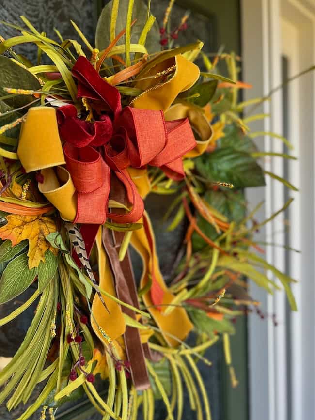 A close-up of a fall wreath decorated with yellow, orange, and red ribbon bows, surrounded by green leaves and artificial foliage, hanging on a dark green front door for a classic home look.