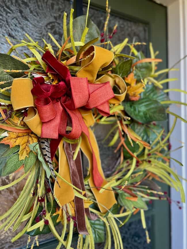 A decorative fall wreath with green leaves, yellow and orange foliage, and a large bow made of mustard yellow, burgundy, and red ribbons adds a classic look to a dark green front door.