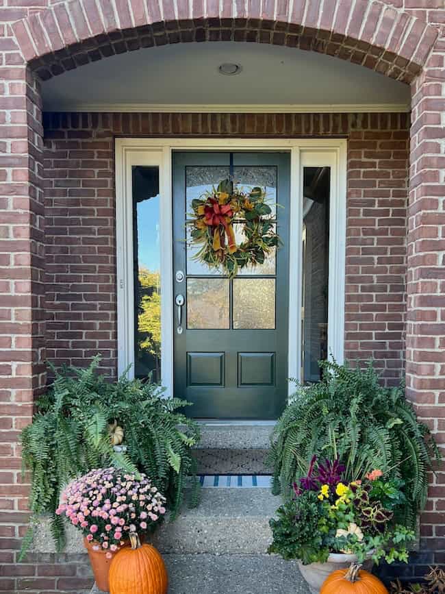 A dark green front door decorated with a fall wreath is flanked by large ferns, colorful flowers, and pumpkins, all set against a brick house exterior for a classic home look.