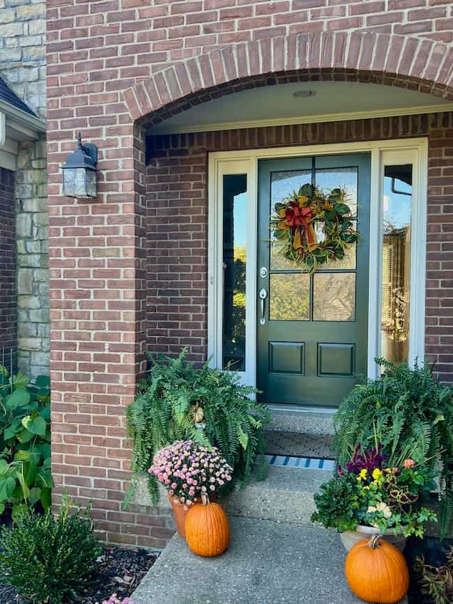 A dark green front door with a fall wreath is framed by a brick archway. Ferns, potted flowers, and three pumpkins are arranged on the steps, creating an autumn-themed porch display with a classic look.