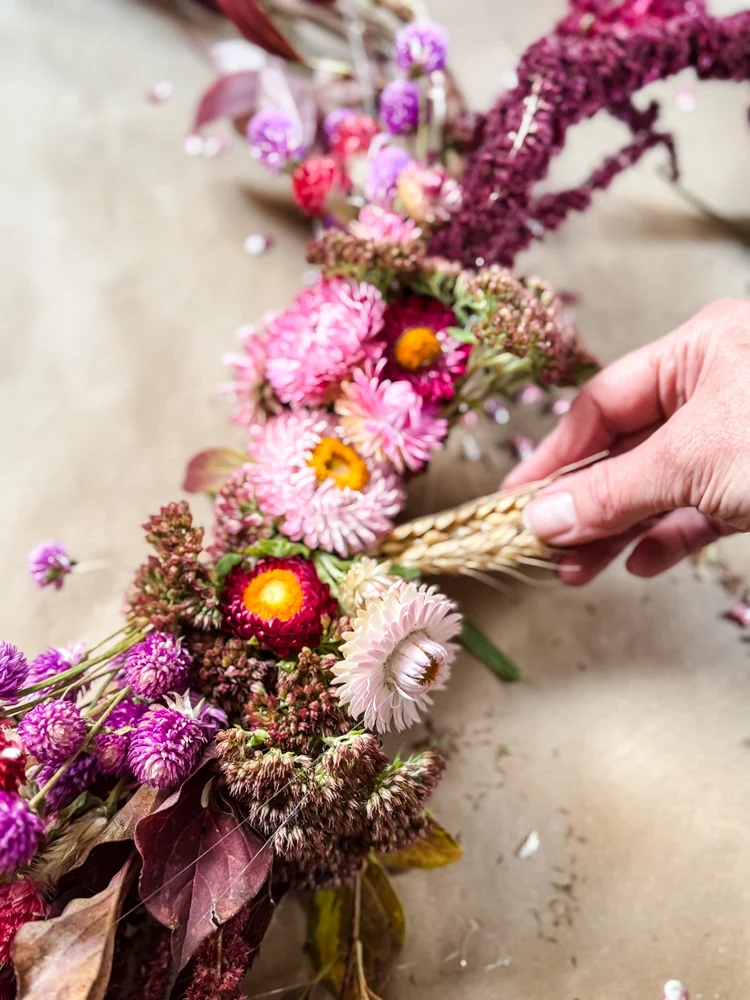 A hand arranges a floral wreath made of dried pink, purple, and red flowers, as well as wheat stalks, on a neutral surface—perfect weekend inspiration for your favorite things.