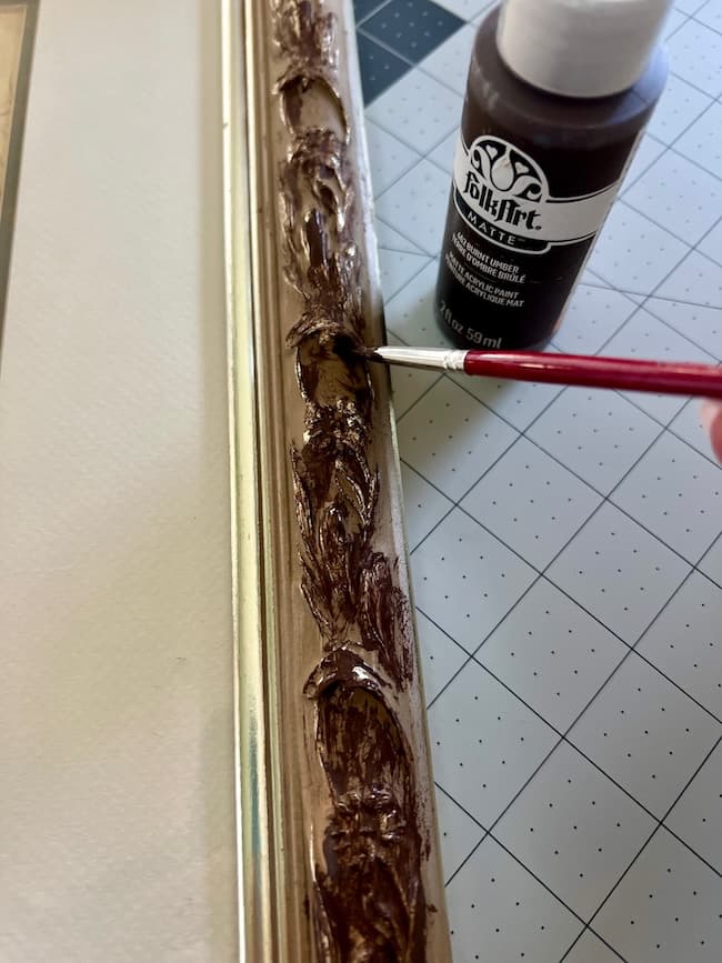 A hand uses a small paintbrush to apply dark brown FolkArt acrylic paint to the ornate edge of a gold picture frame, with a cutting mat underneath—perfect for showcasing your favorite free fine art prints.