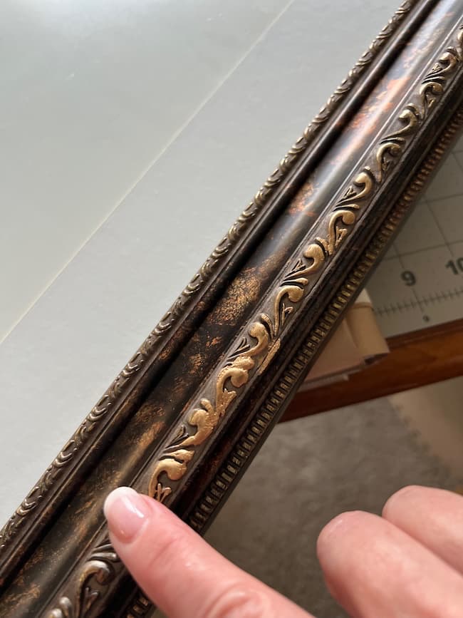 A hand with a finger pointing to an ornate, dark-colored picture frame featuring a decorative leaf pattern near a glass-topped table, part of a ruler visible in the background—perfect for showcasing your free fine art prints.