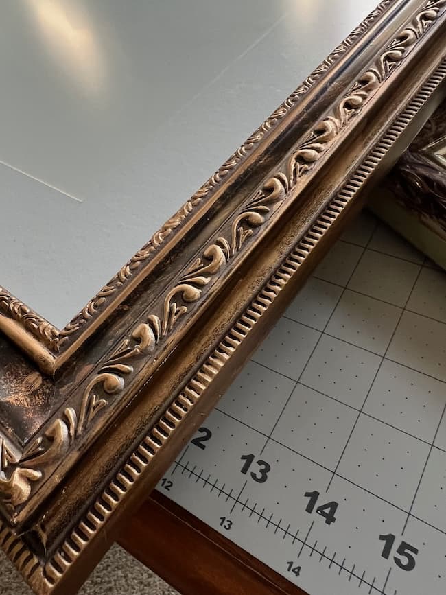 A close-up of an ornate, gold-colored picture frame resting on a gridded cutting mat with inch measurements. The decorative, leaf-like embossed pattern adds elegance—perfect for showcasing your free fine art prints.