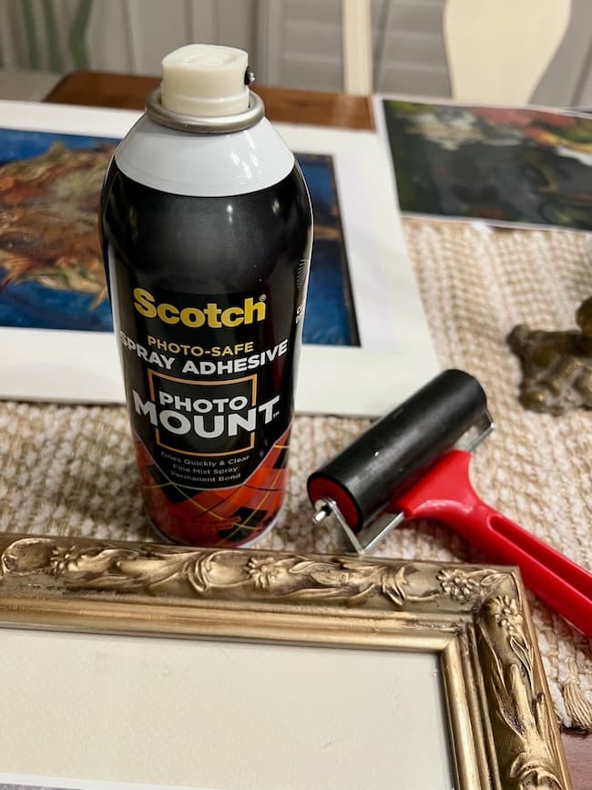 A can of Scotch Photo Mount spray adhesive and a red-handled roller are placed on a table next to an ornate gold picture frame, with free fine art prints and other artwork visible in the background.