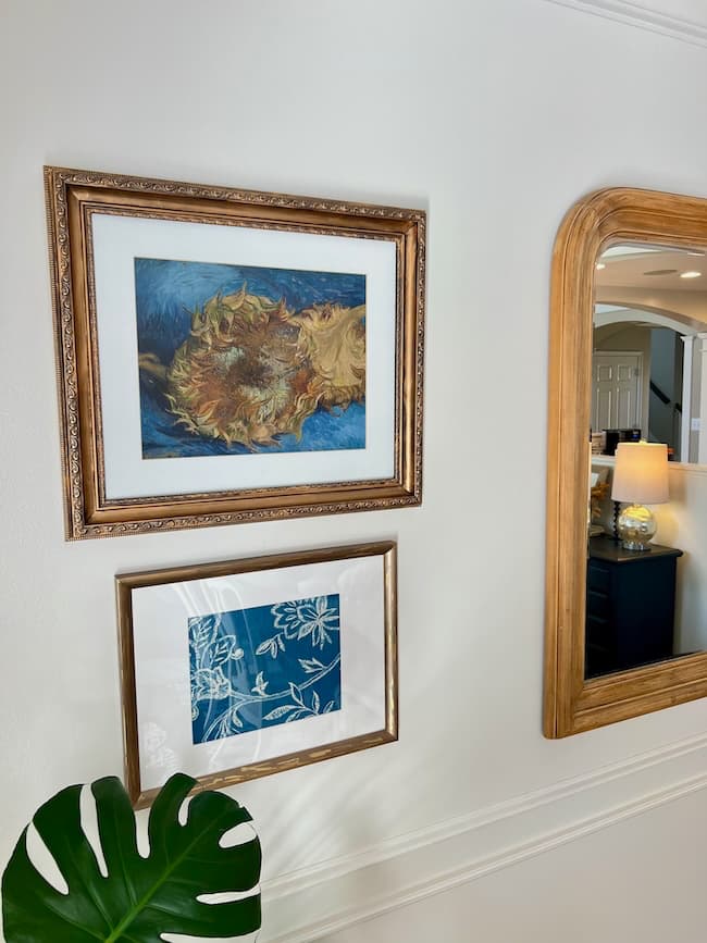 Framed artwork and a large mirror hang on a white wall above a green monstera leaf. The top artwork depicts a swirling abstract scene, while the lower piece features white floral outlines on a blue background.