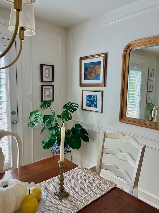 A bright dining room with a wooden table, white chairs, a gold candle holder with a lit candle, a bowl of fruit, green plant in a blue pot, wall art, a large mirror, and sunlight coming through window blinds.