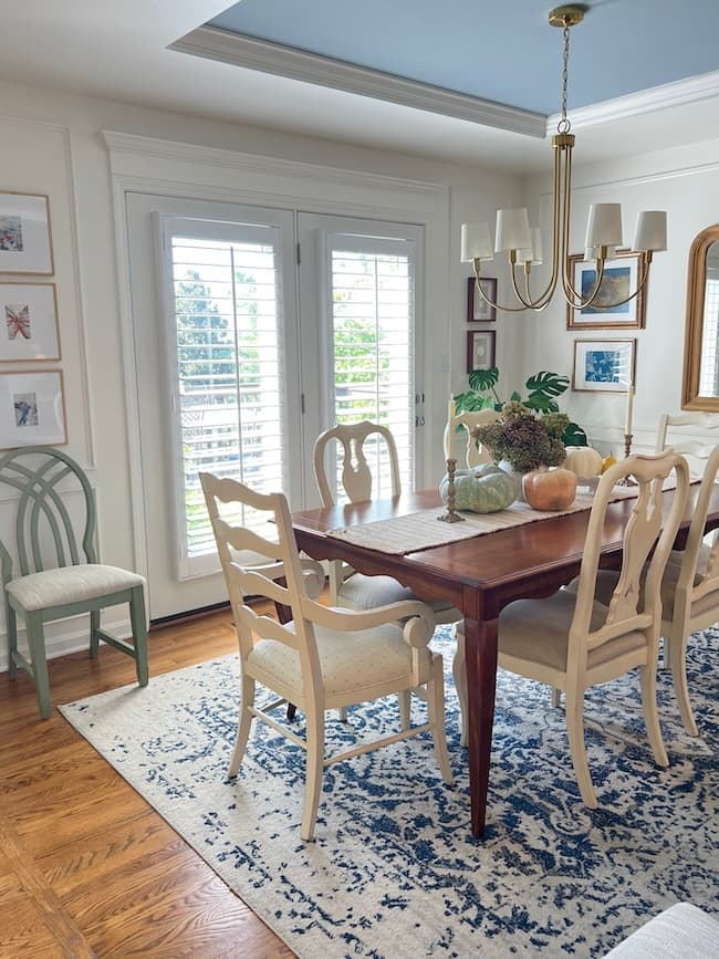 A bright dining room with a wooden table, six cream-colored chairs, a blue patterned rug, a modern chandelier, a gallery wall of art, and French doors with white shutters letting in natural light.