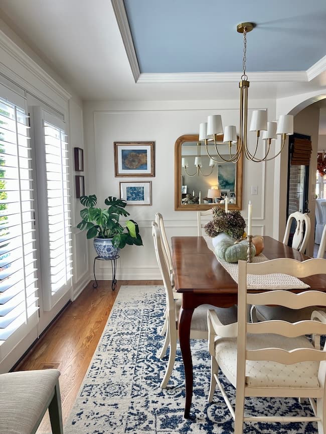Bright dining room with a wooden table, white chairs, a blue and white rug, chandelier, potted plant, wall art, and a mirror reflecting decor; sunlight streams in through large window shutters.