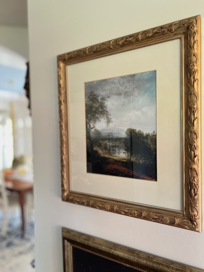 A framed landscape painting with a gold ornate frame hangs on a wall. The artwork depicts trees, a river, and distant hills under a cloudy sky. A sunlit table and chairs are visible in the background.