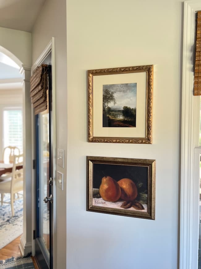 Two framed paintings hang on a light-colored wall; the top is a landscape, and the bottom depicts two oranges. A dining room with a table and chairs is visible through an open doorway.