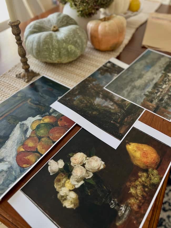 Four printed art reproductions featuring still lifes and landscapes are spread across a wooden table decorated with pumpkins, gourds, and a textured runner.