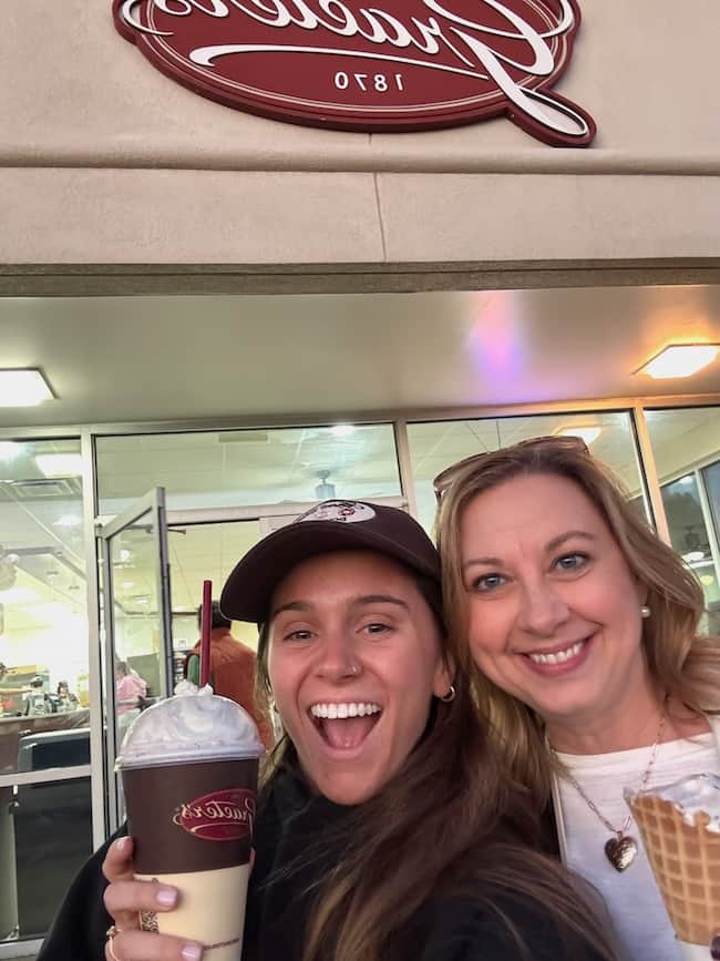 Two smiling women pose outside an ice cream shop, one holding a milkshake and the other an ice cream cone. The shop’s cheerful vibe is enhanced by charming home touches in its decor, making the scene feel both lively and inviting.