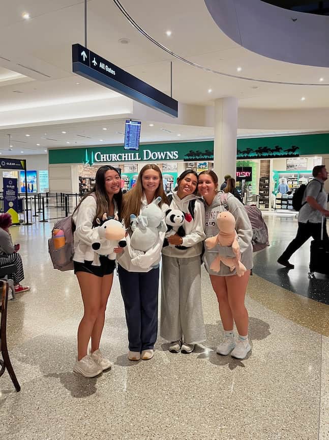 Four smiling young women stand together in an airport terminal holding stuffed animals. Signs, including one for “All Gates,” and a “Churchill Downs” store are visible in the background, capturing fresh ideas and October inspiration. Travelers walk nearby.