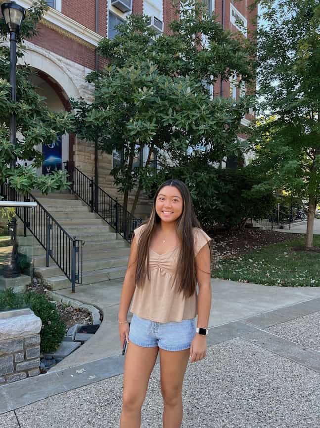 A young woman with long brown hair smiles at the camera while standing outside a brick building with stairs and greenery. She wears a tan top, denim shorts, and a smartwatch—perfect October style and fresh inspiration for Seven on Saturday.