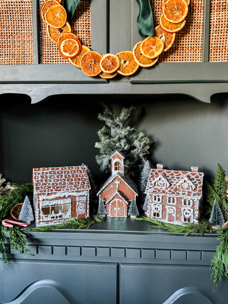 A display of three gingerbread houses with white icing sits on greenery, flanked by mini pine trees. Above, a holiday wreath with dried orange slices hangs on a dark wooden shelf, creating a warm home vibe and beautiful finds for Seven on Saturday.