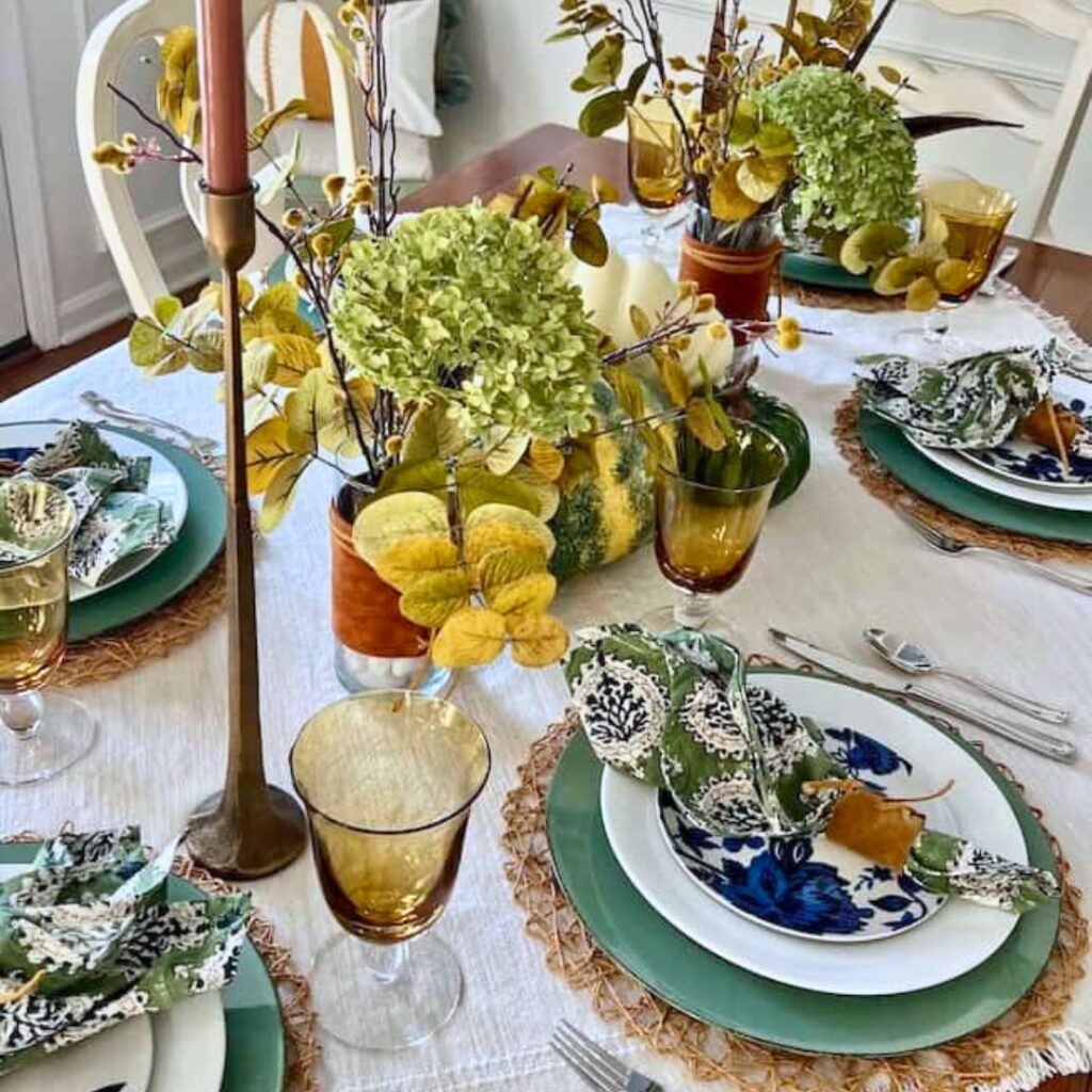 A dining table set for four with green and blue floral plates, green napkins, amber glasses, and woven placemats. The centerpiece of hydrangeas and yellow foliage offers perfect Thanksgiving tablescape ideas with an autumnal theme.