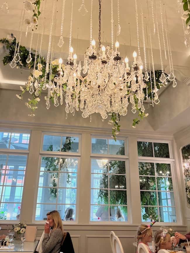 A crystal chandelier with hanging beads and white flowers decorates a bright, elegant room with large windows. People sit at tables below, enjoying a meal in a stylish café filled with beautiful finds and a warm home atmosphere.