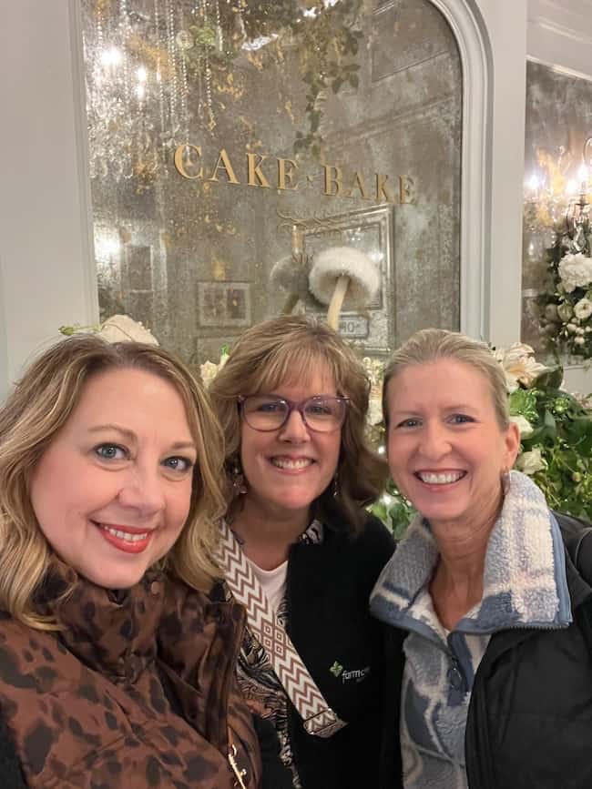 Three women smiling and posing together indoors in front of a decorated window that says "Cake Bake," surrounded by flowers and soft lighting, create a scene full of beautiful finds and warm home vibes.