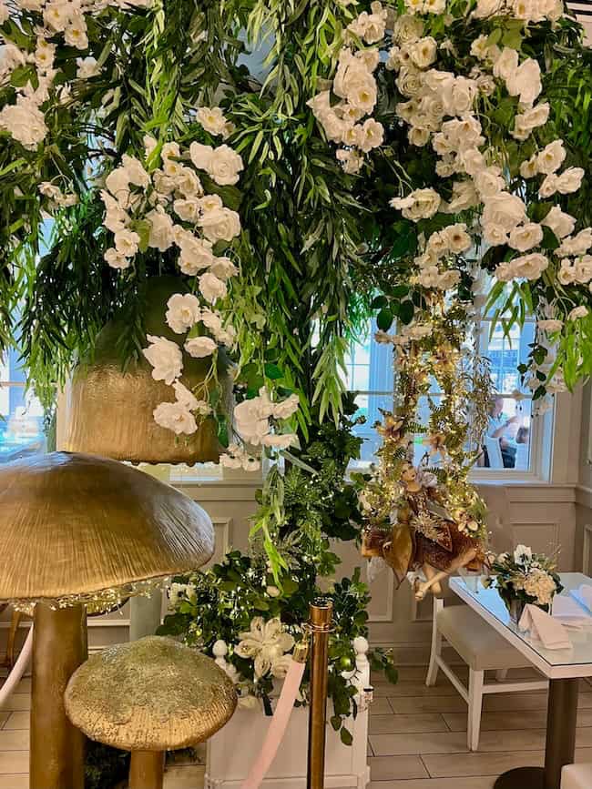 A restaurant interior decorated with large artificial mushrooms, cascading white flowers, and lush green foliage creates a whimsical, garden-like atmosphere—perfect for discovering beautiful finds reminiscent of a Warm Home. White tables and chairs are seen in the background.