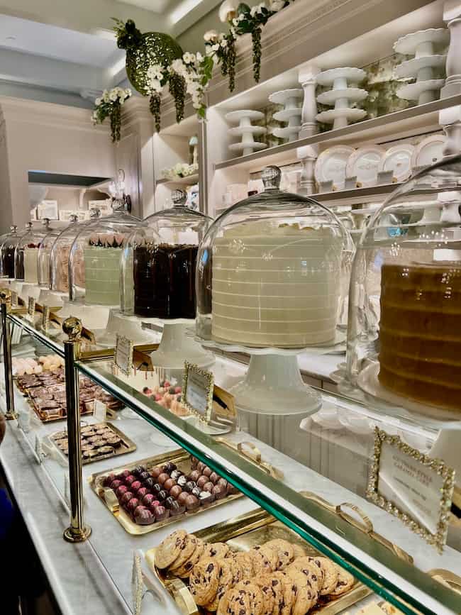 A bakery display with several large cakes under glass domes, trays of assorted pastries, cookies, and chocolates in front, and shelves of white dishes and decor in the background—truly a Warm Home filled with Beautiful Finds.