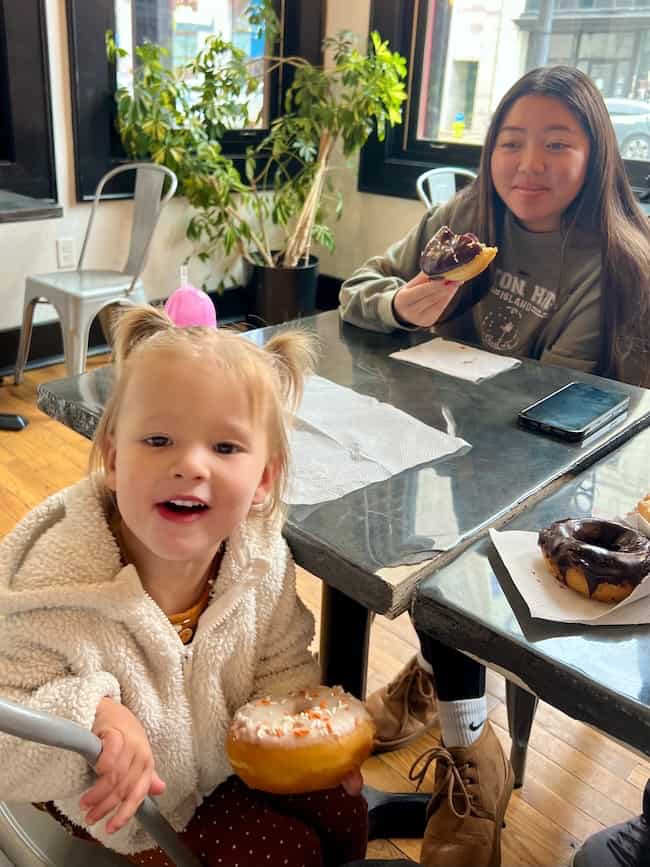 A young girl smiles while holding a donut, sitting at a café table with a woman who is also enjoying these beautiful finds. The table has more donuts and a phone on it, with plants and windows giving the space a warm home feel.