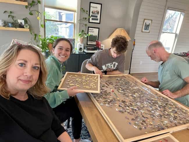 Four people sit around a wooden table working on a jigsaw puzzle. One woman smiles at the camera, another holds up a puzzle board, while two others focus on pieces—creating warm home memories among plants and pictures in the background.