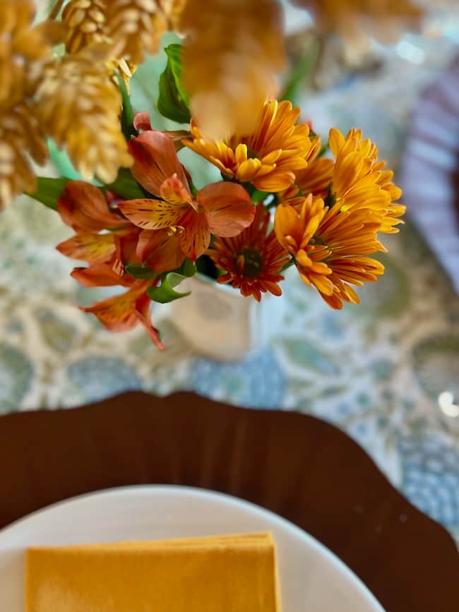 A close-up of an autumn-themed flower arrangement with orange and yellow blooms in a vase on a patterned tablecloth, creating a charming fall style Thanksgiving tablescape on a small table next to a white plate with a folded mustard-yellow napkin.
