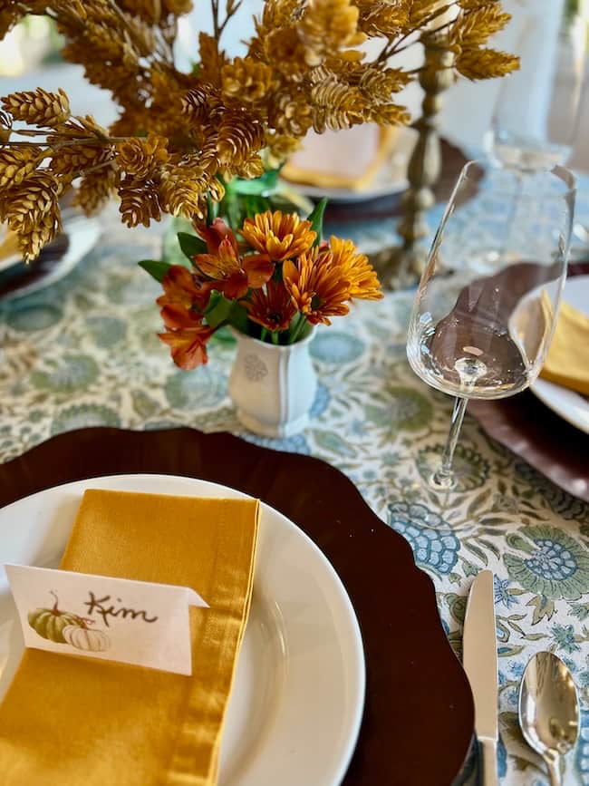 A fall style Thanksgiving tablescape on a small table features a white plate, gold napkin, and "Kim" name card. Autumn flowers in a vase and a wine glass rest on a patterned tablecloth with brown and gold accents.