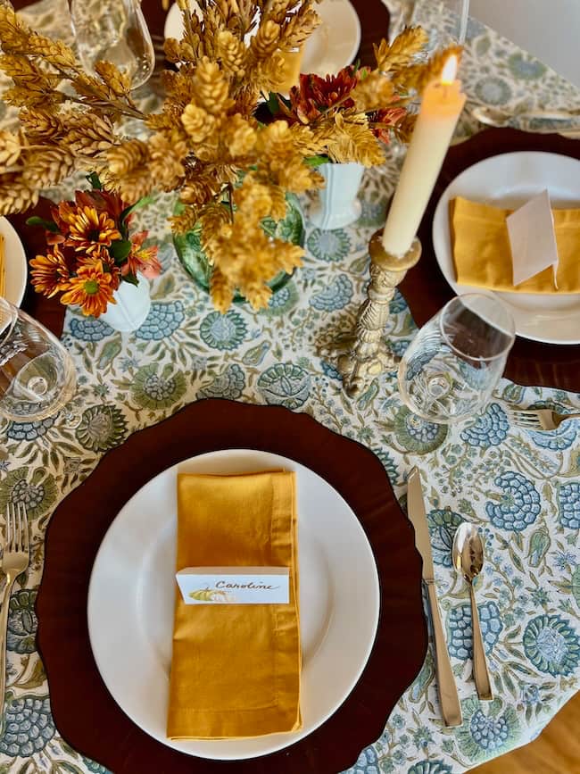A decorated small table showcases a fall style Thanksgiving tablescape with a floral tablecloth, gold utensils, wine glasses, yellow flowers, a lit candle, and plates set with gold napkins and name cards.