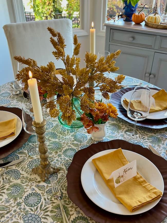 A Thanksgiving tablescape features a small table with yellow napkins, white plates on dark chargers, a floral tablecloth, gold candlesticks, and autumn foliage as the centerpiece. A place card with "Kim" rests on one plate, completing the fall style.