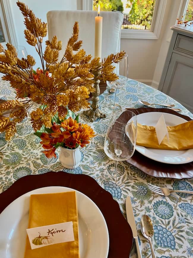 A round small table set for a meal with autumn-themed decorations creates a cozy Thanksgiving tablescape—gold napkins, orange flowers, a brown charger plate, and a handwritten place card reading "Kim." A lit candle glows in the background.