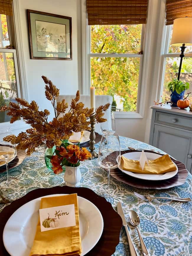 A small table is set for a meal with autumn-themed decor, featuring a floral tablecloth, yellow napkins, white plates, wine glasses, and a dried plant centerpiece in a green vase—a cozy Thanksgiving tablescape bathed in sunlight.