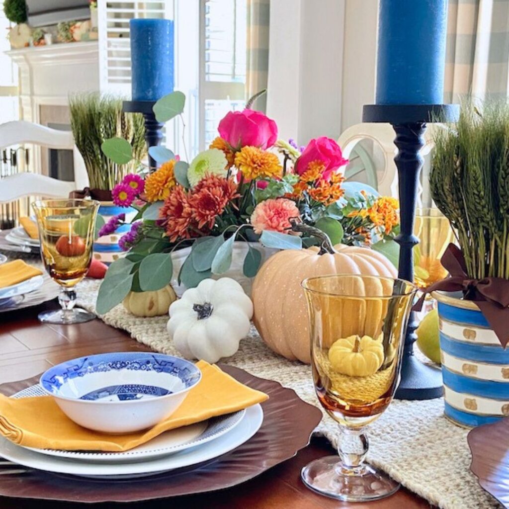 A fall-themed holiday table with blue candles, yellow glasses, blue and white bowls, orange napkins, pumpkins, gourds, and a centerpiece of colorful flowers and greenery—perfect for Thanksgiving décor.