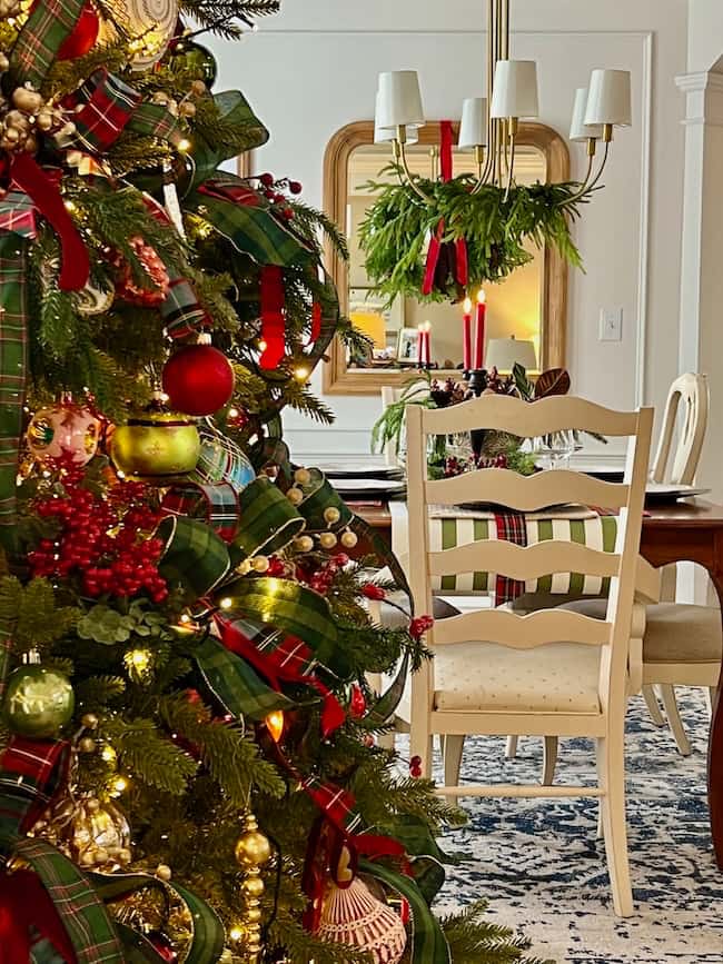A decorated Christmas tree with red and green ornaments stands in the foreground; in the background, a dining table with white chairs, greenery, and red candles is visible, creating a festive holiday atmosphere.