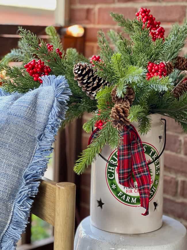 A festive arrangement with pine branches, red berries, and pinecones sits in a white metal container labeled "Christmas Trees." A red plaid ribbon is tied around the container; a blue fringed blanket is draped nearby.