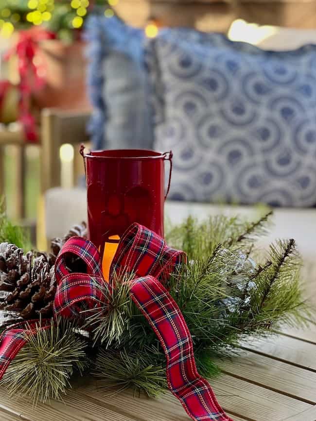 A festive centerpiece with pine branches, pinecones, a red plaid ribbon, and a red lantern sits on a wooden table in front of a couch with blue patterned pillows.