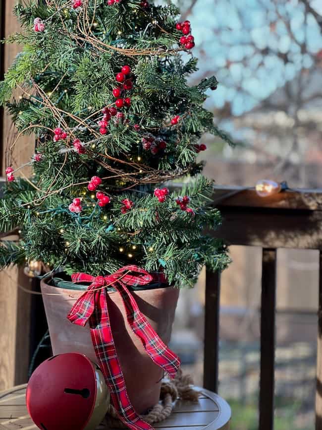 A small potted Christmas tree decorated with red berries, gold lights, and a red plaid ribbon sits on a round outdoor table next to a large red jingle bell ornament.