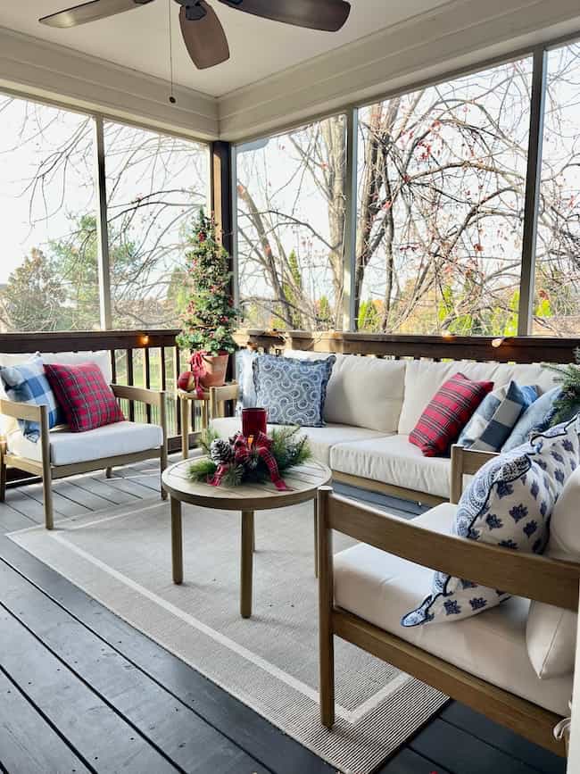 A cozy screened porch with a white sofa, two armchairs, and a round table decorated for the holidays. Red and plaid pillows, a small Christmas tree, and greenery with candles add a festive touch.