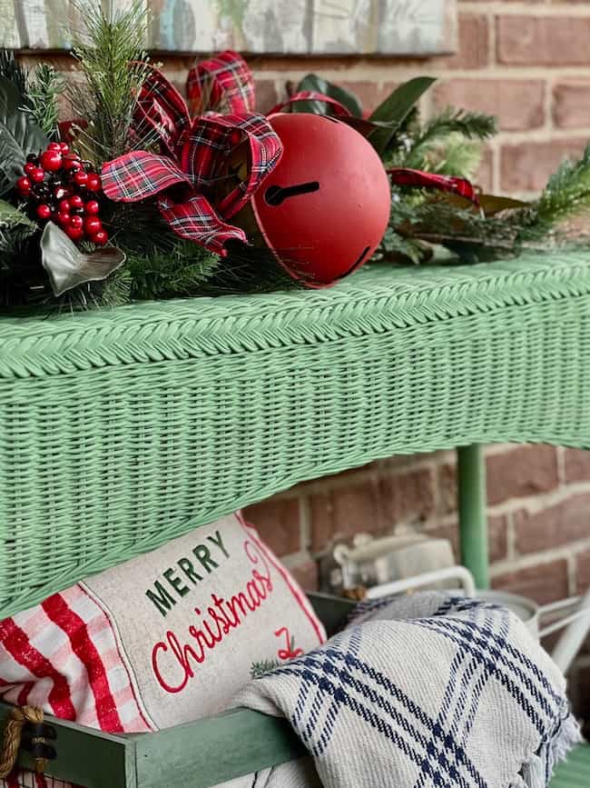 A green wicker table decorated with pine branches, red berries, a plaid ribbon, and a large red jingle bell. Below, a pillow reads "Merry Christmas" and a blue-and-white checkered blanket is draped nearby.