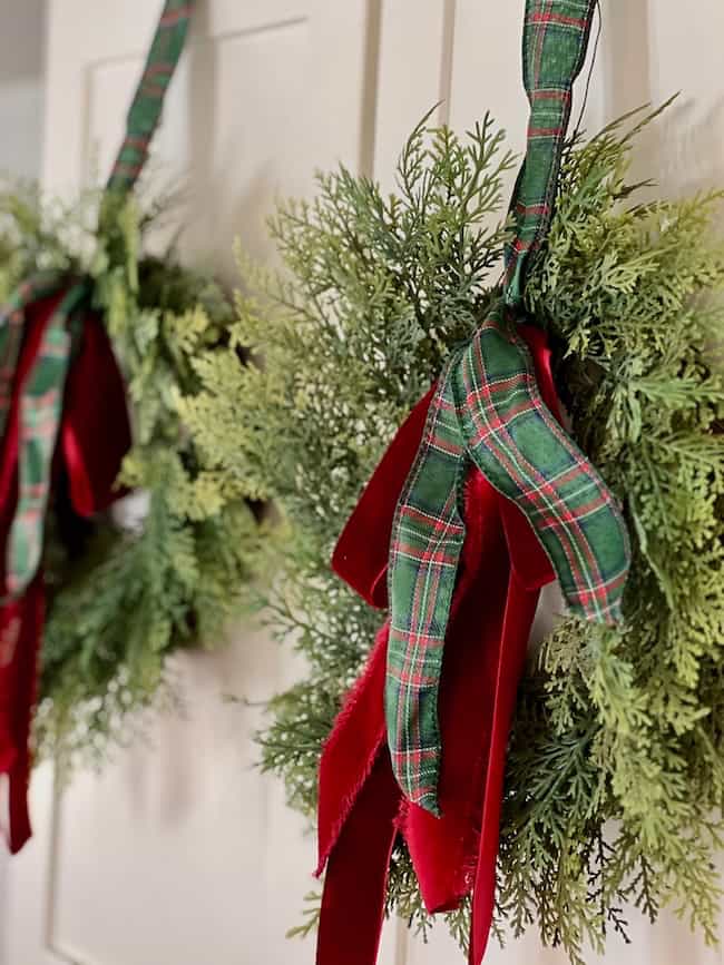 A close-up of two green holiday wreaths hanging on a door, decorated with red velvet and green plaid ribbon bows.