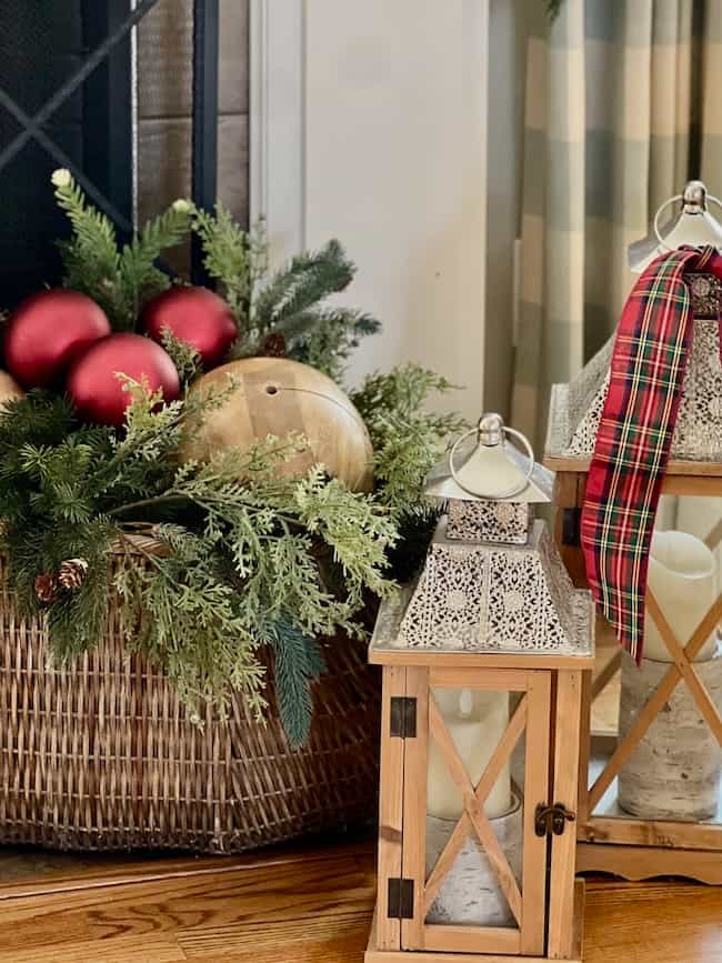 A wicker basket with evergreen branches, red ornaments, and a wooden ball sits beside two decorative lanterns, one adorned with a red plaid ribbon, on a wooden floor near a fireplace.