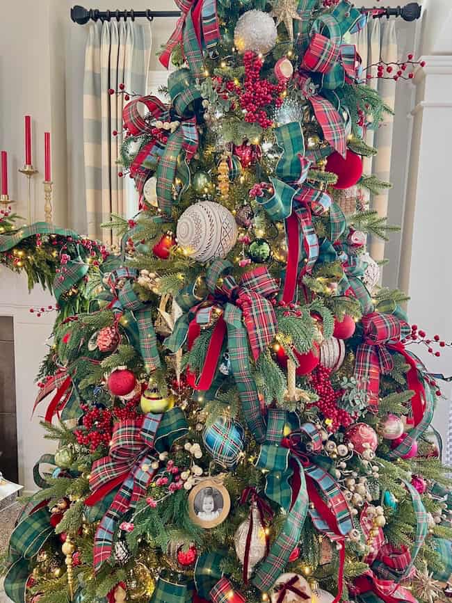 A Christmas tree decorated with plaid ribbons, red berries, ornaments, and framed photos. There are red candles on a mantel in the background and cream-colored curtains on the window.