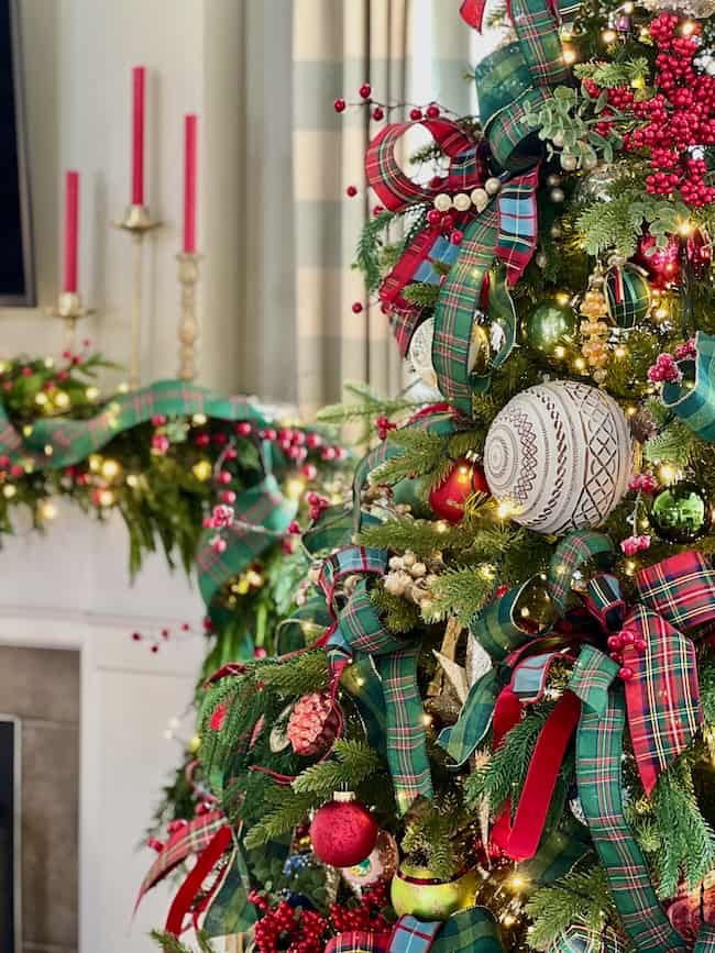 A Christmas tree decorated with plaid ribbons, red and green ornaments, and gold accents stands next to a mantel adorned with garland and red candles, creating a festive holiday scene.