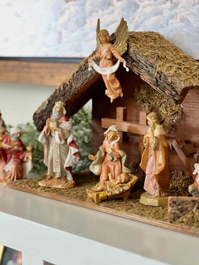 A nativity scene with figurines of baby Jesus in a manger, Mary, Joseph, the three wise men, an angel above, and a rustic stable setting with moss on the roof.