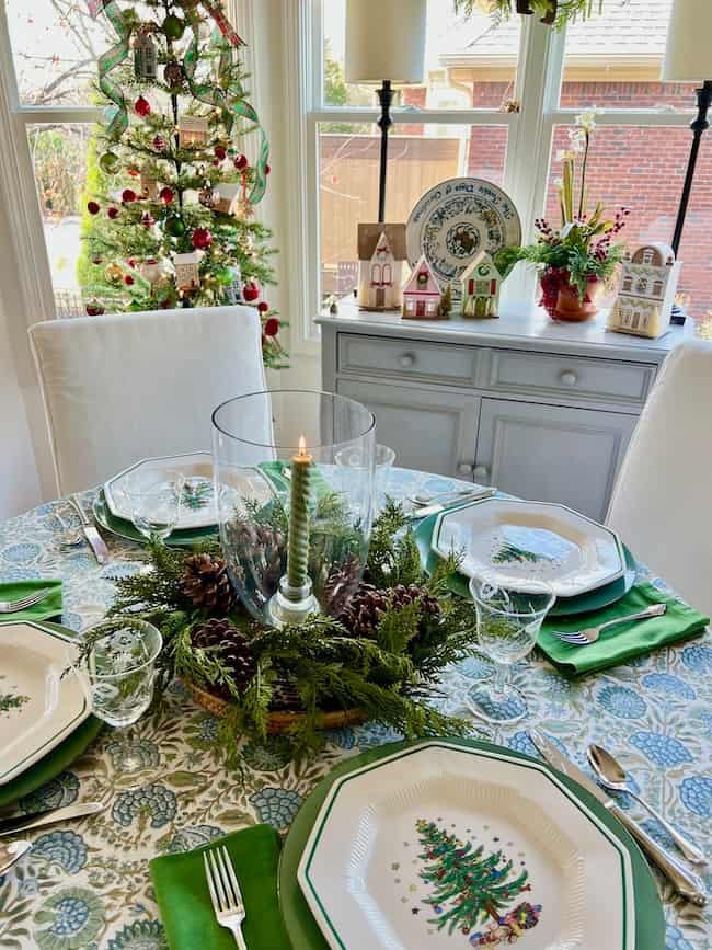 A festive dining table set for four with Christmas-themed plates, green napkins, and a candle centerpiece surrounded by greenery. In the background, a decorated Christmas tree and holiday decorations are visible.
