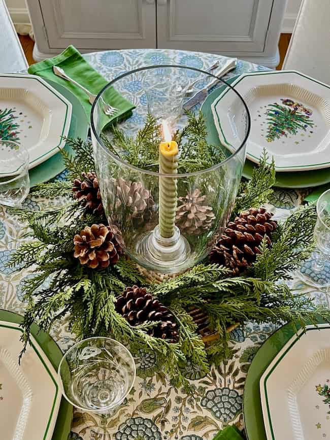 A festive table centerpiece with a lit striped candle in a glass holder, surrounded by pinecones and evergreen branches. The table is set with plates, green napkins, and clear glasses on a patterned tablecloth.