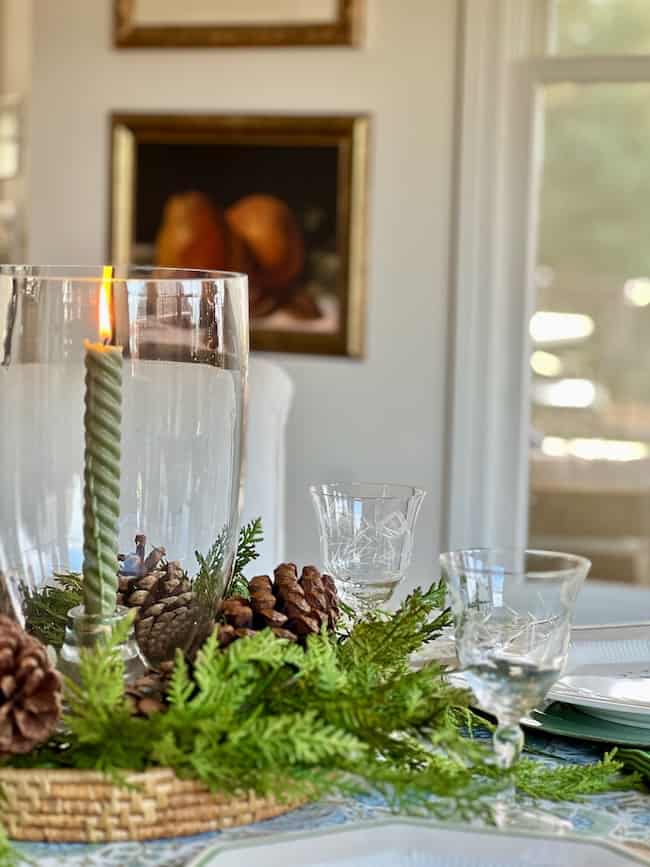 A close-up of a dining table centerpiece featuring a green candle in a glass holder, surrounded by pinecones and evergreen branches. Elegant glassware and plates are set on the table in a bright room.