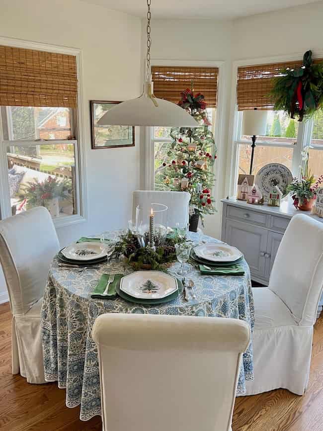 A round dining table with a blue and white tablecloth is set for four, each place with a white plate featuring a winter tree, green napkins, and glassware. Christmas decorations and a tree are seen in the cozy, sunlit room.